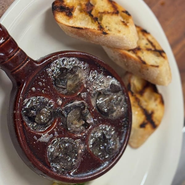 Escargot in a brown ceramic dish with six compartments, served with two slices of grilled bread.