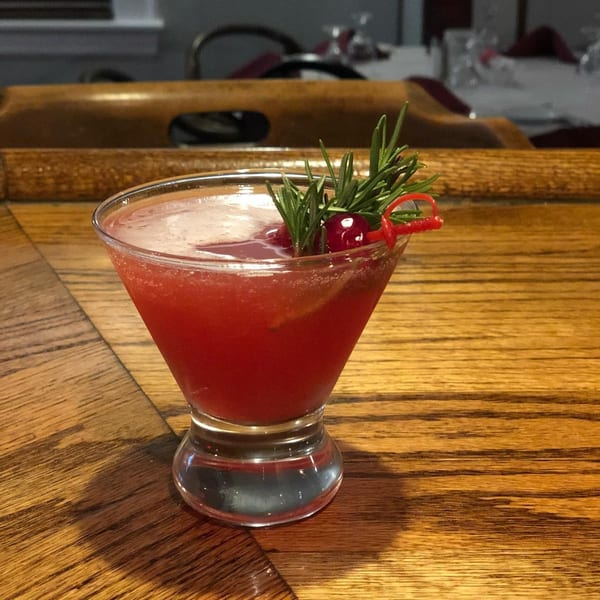 A red cocktail in a martini glass, garnished with rosemary and cranberries, on a wooden table.