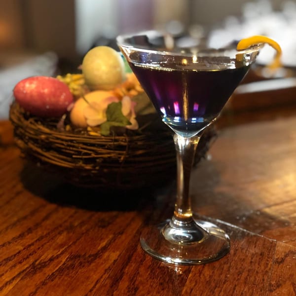 A purple cocktail with a lemon twist next to a decorative nest with colorful eggs on a wooden table.
