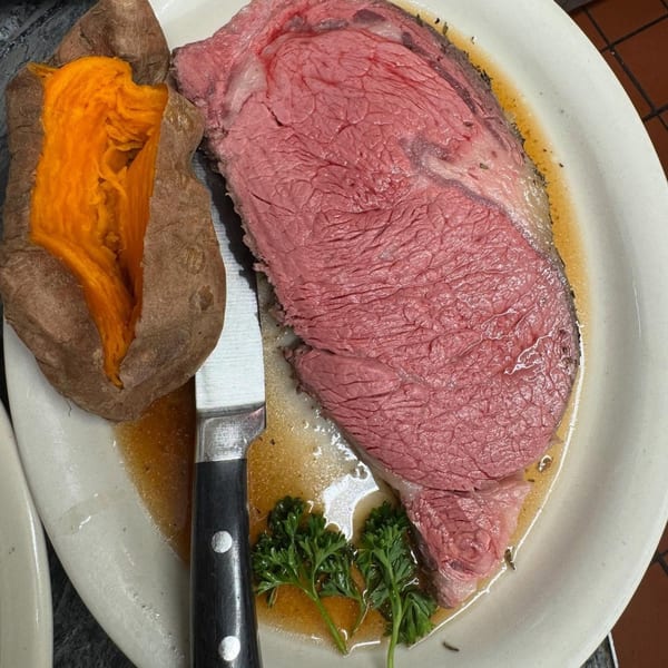 A Coach & Four prime rib steak with a baked sweet potato, knife, and parsley on a white plate.