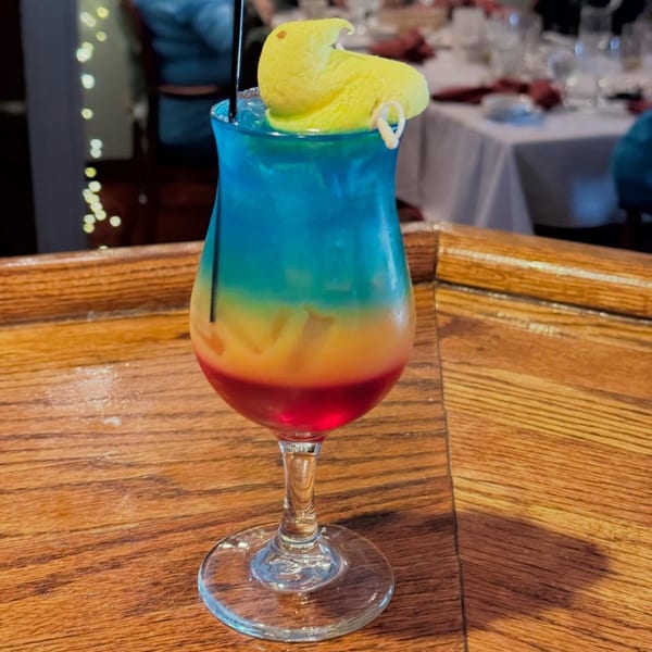 A colorful layered cocktail with a yellow Peep candy garnish sits on a wooden bar.